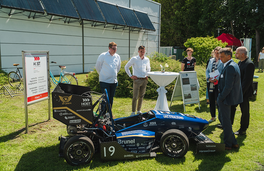 Personen in Anzügen schauen sich den blauen Formel-Student-Rennwagen des Baltic Racing Teams im Sonnenschein an