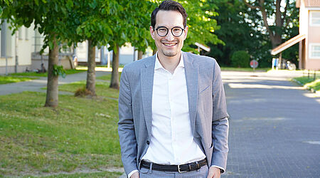 Ein lächelnder Mann im Anzug steht vor einer Baum-Allee auf dem Campus: Kanzler Fabian Berger
