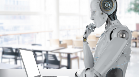 Ein Roboter, eine künstliche Intelligenz, sitzt nachdenklich vor einem Laptop
