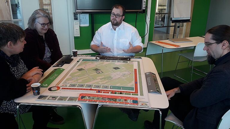 Prof. Dr. Johannes Gulden (r.) und Romy Sommer (l.) bekommen in Groningen an der Hanze eines der Serious Games vorgestellt: Vor vier Menschen auf dem Tisch liegt eine Art Brettspiel, großformatig mit vielen eingezeichneten Grünflächen auf einer Art Karte.