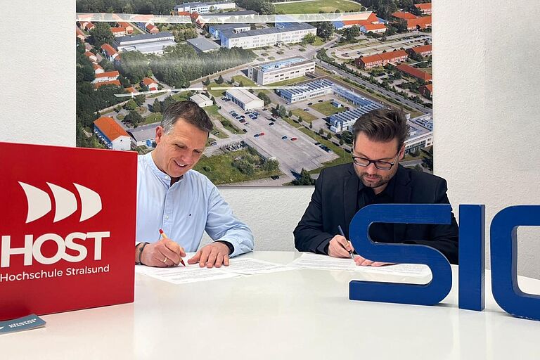 Zwei Männer unterschreiben jeweils ein Papier an einem Schreibtisch sitzend