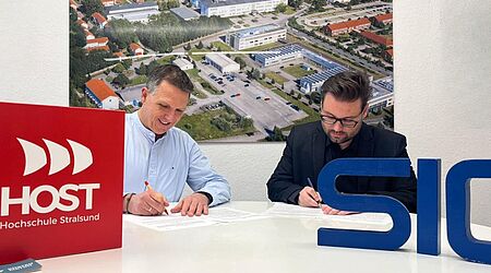 Zwei Männer unterschreiben jeweils ein Papier an einem Schreibtisch sitzend