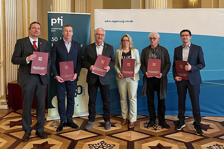 Eine Gruppe aus sechs Personen, fünf Männern und einer Frau, sie halten je ein A4-Blatt in der Hand, einen Fördermittelbescheid, es ist das Team hinter AutoPasture: Prof. Mark Vehse (Hochschule Stralsund, v.l.), Prof. Tobias Hillmann (Hochschule Neubrandenburg), Dr. Jan Langbein (Forschungsinstitut für Nutztierbiologie), Prof.in Sandra Rose (Universität Rostock), Dr. Jürgen Müller (Universität Rostock), Martin Priesel (Forschungsverbund MV) 