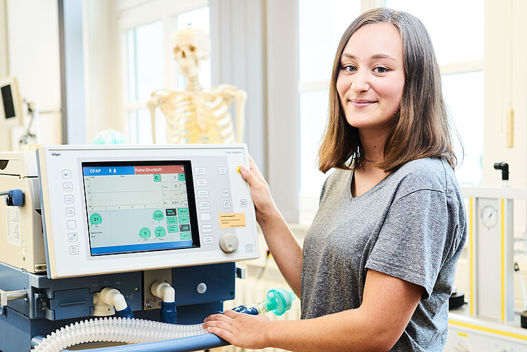 Eine Junge Frau steht neben einem Computer-Bildschirm im Hintergrund ist ein medizinisches Skelett zu sehen
