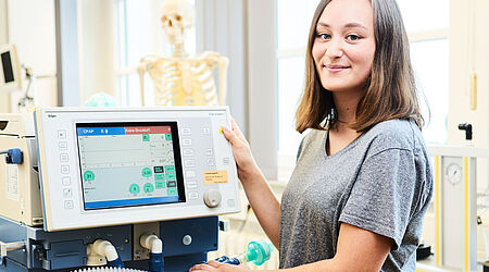 Eine Junge Frau steht neben einem Computer-Bildschirm im Hintergrund ist ein medizinisches Skelett zu sehen
