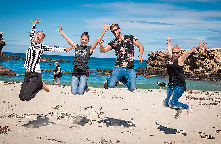 4 springende Studierende in sommerlicher Kleidung am Strand
