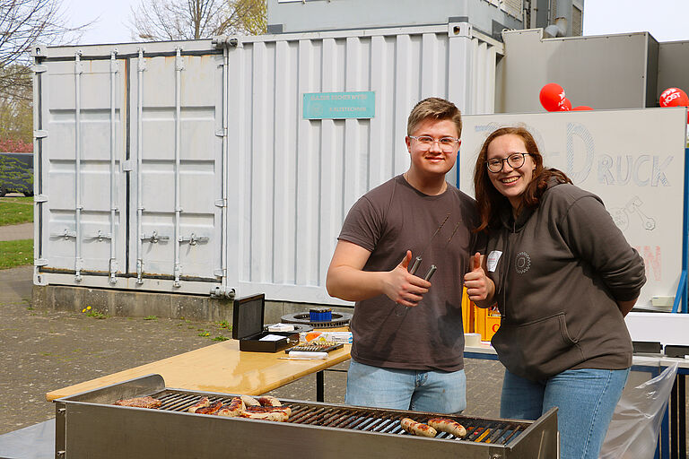 Zwei Studierende stehen fröhlich vor einem Grill