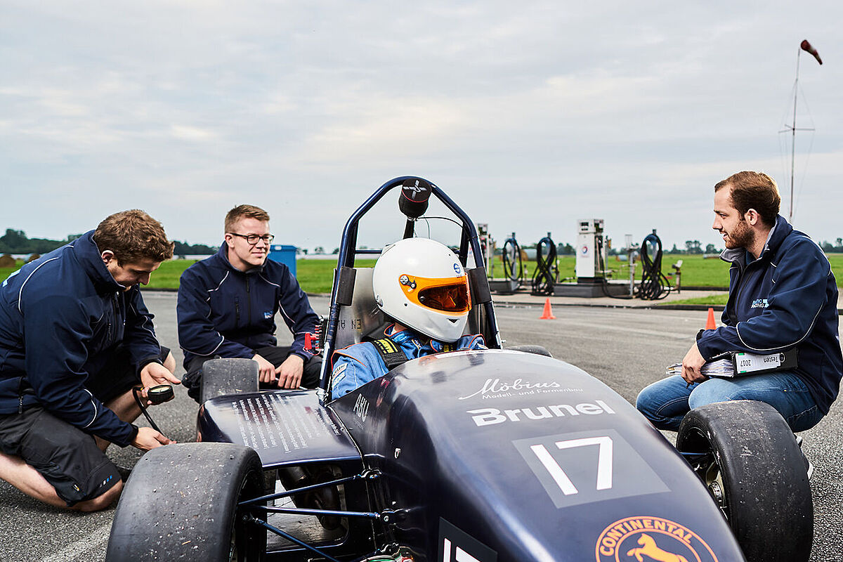 Kniende und stehende Studierende in Kontakt mit dem Fahrer in einem Student Racing Rennwagen auf einer Rennstrecke