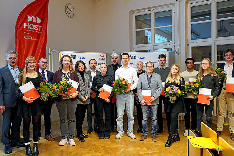 16 Frauen und Männer unterschiedlichen Alters, in Anzügen und Blaisern, in einem Hörsaal der Hochschule. Die Studierenden unterihnen halten Urkunden in die Kamera.