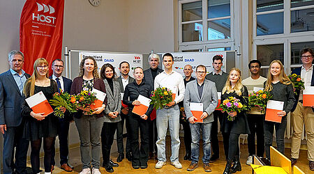 16 Frauen und Männer unterschiedlichen Alters, in Anzügen und Blaisern, in einem Hörsaal der Hochschule. Die Studierenden unterihnen halten Urkunden in die Kamera.
