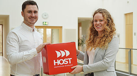 2 Personen mit Hemd und Blaiser halten großen roten Würfel mit HOST-Logo in die Kamera