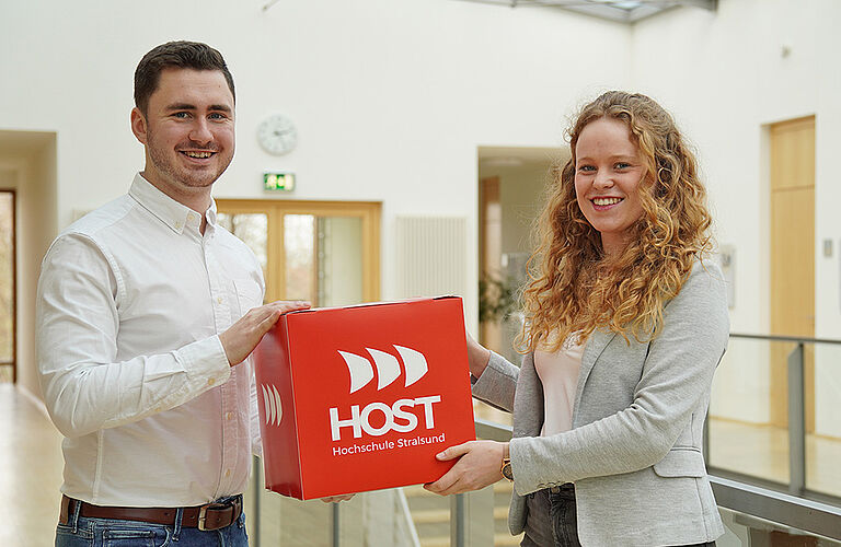 2 Personen mit Hemd und Blaiser halten großen roten Würfel mit HOST-Logo in die Kamera