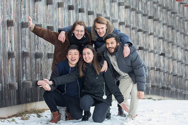 eine Gruppe von fünf jungen Menschen posiert für ein Foto, teils hockend an einer hölzernen Wand im Schnee