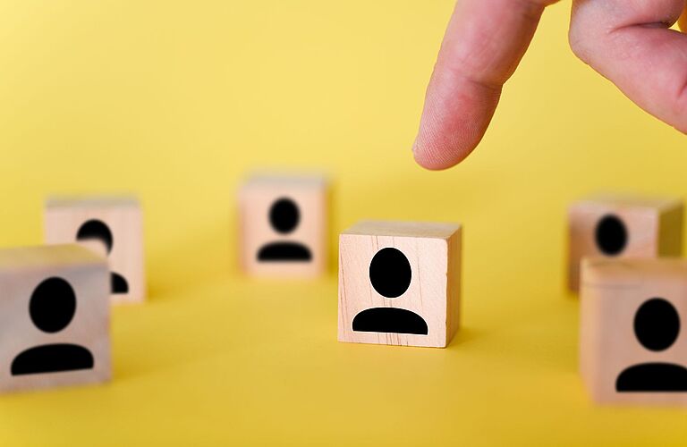Ein gelbes Bild auf dem kleine Holzwürfel mit schemenhaften schwarzen Figuren abgebildet sind und ein Finger, der auf einen weist