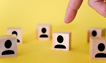 Ein gelbes Bild auf dem kleine Holzwürfel mit schemenhaften schwarzen Figuren abgebildet sind und ein Finger, der auf einen weist