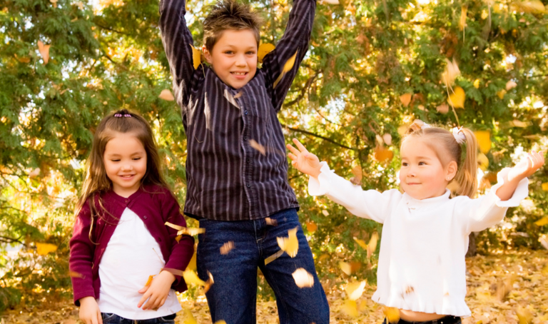 Drei Kinder, zwei kleine Mädchen und ein etwa 10-jähriger Junge, spielen mit Laub, das sie hochwerfen.