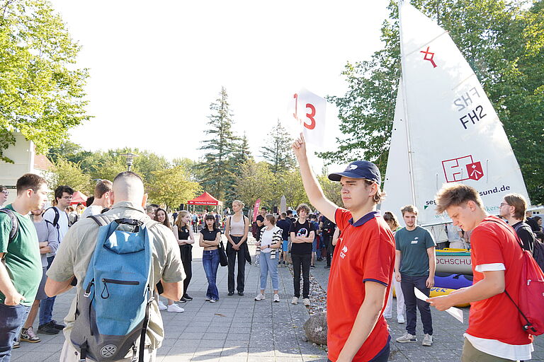 Ein junger Mann im roten Shirt hält einen Zettel mit "1.3" hoch, um ihn herum im Halbkreis junge Leute