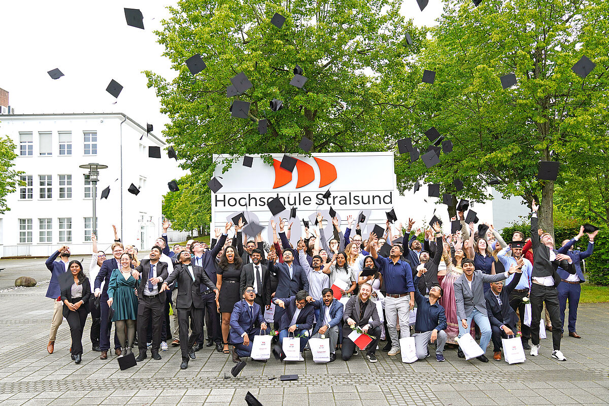 Viele Studierende feiern gemeinsam Ihren Abschluss und werfen Absolventenkappen vor einem großen Holzschild mit der Jahreszahl zweitausendvierundzwanzig in die Luft.