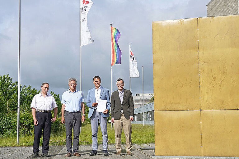 Vier Männer stehen neben einem großen goldenen Würfel, einem Kunstwerk am Bau, vor wehenden Flaggen, darunter die Regenbogenflagge und halten ein Blatt Papier hoch.