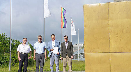 Vier Männer stehen neben einem großen goldenen Würfel, einem Kunstwerk am Bau, vor wehenden Flaggen, darunter die Regenbogenflagge und halten ein Blatt Papier hoch.