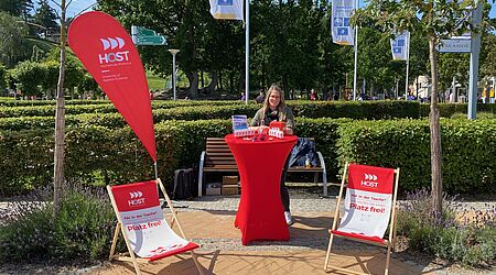Eine junge Frau steht an einem roten Stehtisch der Hochschule Stralsund vorn zwei Liegestühle, neben ihr eine Beach-Flag - alles in Rot-Weiß und mit HOST-Logos.