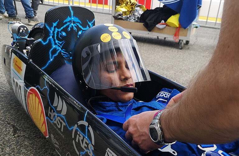 Nahaufnahme: eine junge Rennfahrerin in blauen Rennoverall wird in einem sehr schmalen Rennwagen (ThaiGer7) festgeschnallt