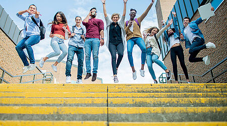 10 Frauen und Männer stehen in sommerlicher Kleidung am oberen Ende einer gelben Treppe und springen in die Luft.