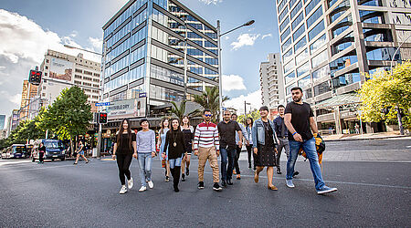 1 Dutzend internationale Studierende in sommerlicher Kleidung überqueren eine Straße mit gläsernen Hochhäusern im Hintergrund