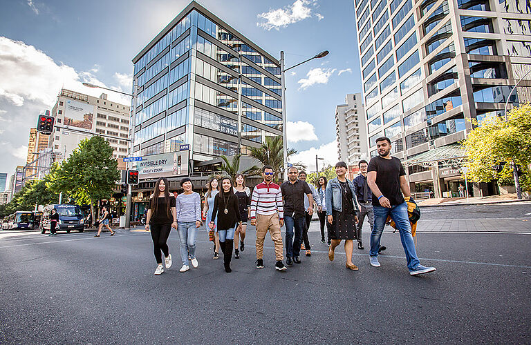 1 Dutzend internationale Studierende in sommerlicher Kleidung überqueren eine Straße mit gläsernen Hochhäusern im Hintergrund