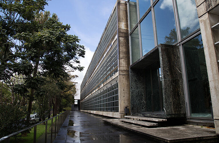 Blick auf einen Gehweg. Links davon ist ein bewaldeter Grünstreifen. Rechts davon befindet sich ein futuristisch aussehendes graues Haus mit anteiligen Glasfassade.