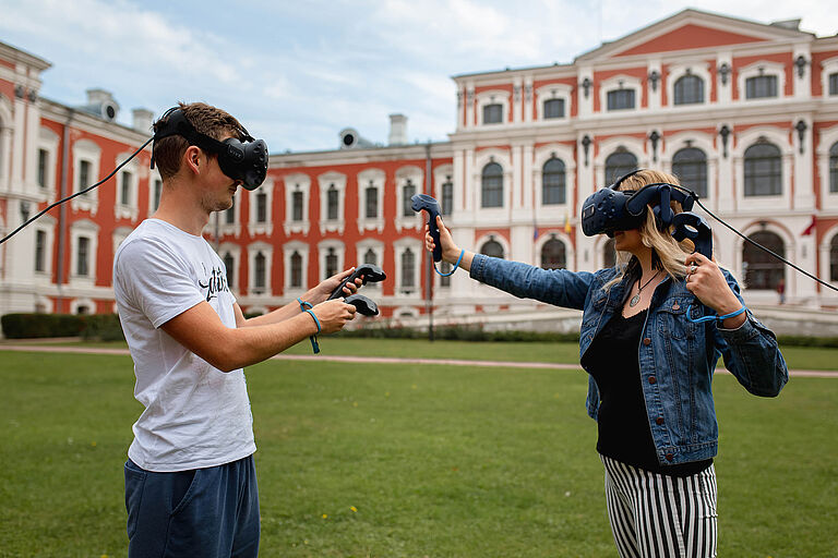 Ein junger Mann und eine junge Frau tragen sich gegenüber stehend große schwarze Virtual-Reality-Brillen und haben Controller in der Hand.