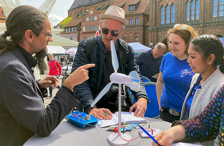 Prof. Johannes Gulden im Gespräch mit 2 Studierenden und einem Mann am Stegtisch mit Modellen von Windkraftanlagen und Solarbooten