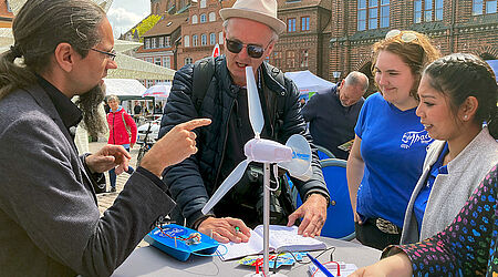 Prof. Johannes Gulden im Gespräch mit 2 Studierenden und einem Mann am Stegtisch mit Modellen von Windkraftanlagen und Solarbooten
