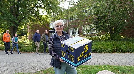 Eine Frau steht mit einer Umzugskiste vor einem Gebäude - die Leiterin vor Bibliothek.