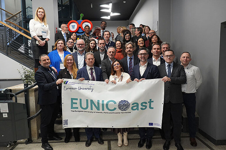 Ca. 3 Dutzend Menschen, in Anzügen und Blaisern, stehen auf einer Treppe, in einem Bürogebäude, und halten ein EUNICoast-Banner in die Kamera.