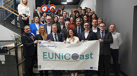 Ca. 3 Dutzend Menschen, in Anzügen und Blaisern, stehen auf einer Treppe, in einem Bürogebäude, und halten ein EUNICoast-Banner in die Kamera.