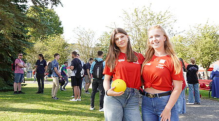 Zwei junge Frauen stehen in roten T-Shirts vor einer Gruppe junger Menschen, den neuen Studierenden