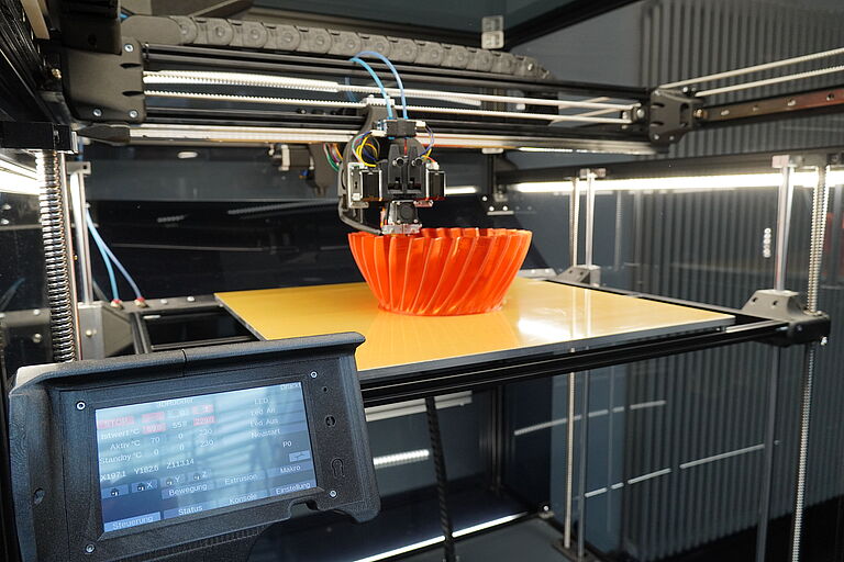 Zu sehen ist ein Blick in den 3D-Drucker und wie gerade Schicht um Schicht Filament auf eine orangefarbene Schale aufgetragen wird. 