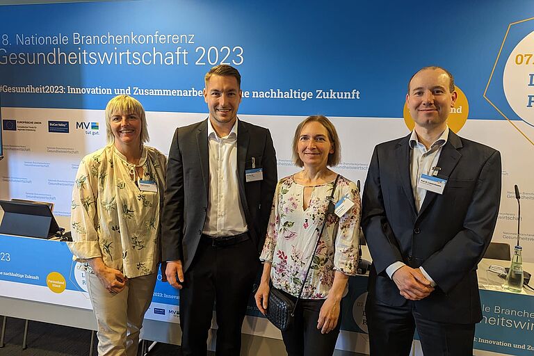 Vier Personen, zwei Männer und zwei Frauen, stehen lächelnd zusammen vor Plakaten vermeintlich einer Messe. 