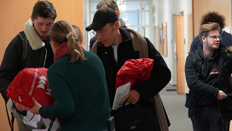 Studierende nehmen vor dem Hörsaal von einer Frau ihre Willkommensbeutel, rote Rucksackbeutel mit HOST-Logo, entgegen.