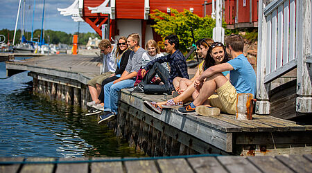 eine Gruppe junger, fröhlicher Menschen sitzt, sommerlich gekleidet, auf einem Steg.