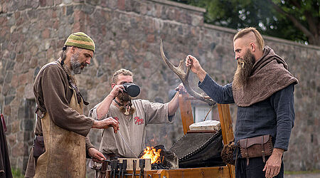 3 Männer in Mittelalterkutte stehen mit Werkzeug an einem Tisch mit Flamme und Aufsätzen