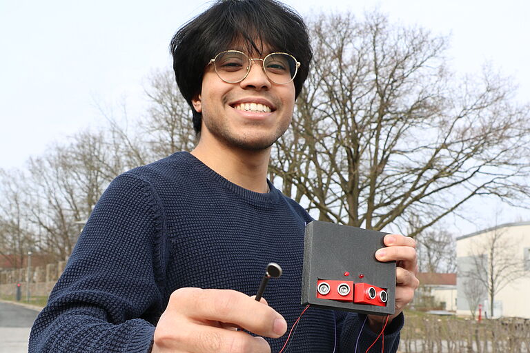 ein lächelnder junger Mann, Mustafa Rahmati, hält einen kleinen grauen Kasten mit vier lautsprecherähnlichen Sensoren und roten Kabeln