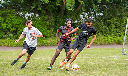 Drei Personen spielen Fußball auf einem grünen Rasen.