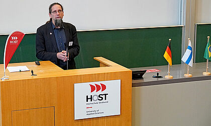 Prof. Johannes Gulden, in schwarzen Anzug, steht an einem Pult mit dem Logo Hochschule Stralsund, vor einer grünen Tafel.