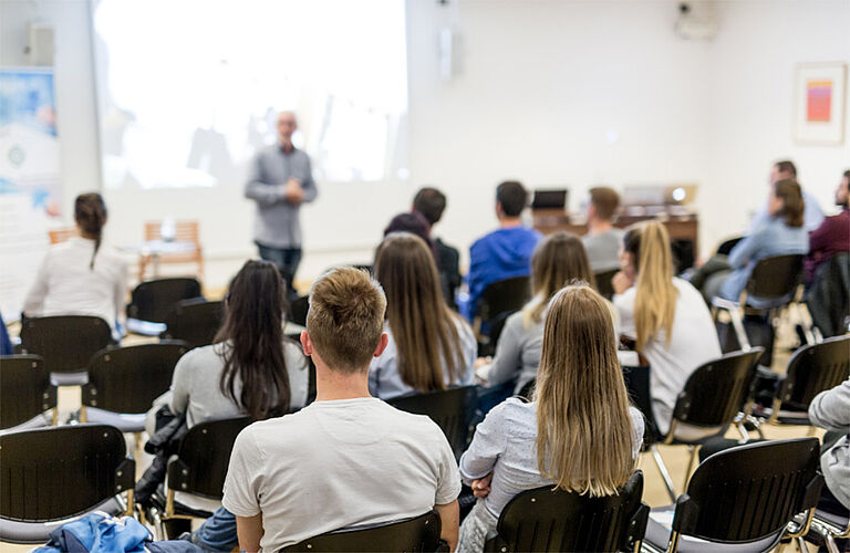 Hörsaal mit besetzten Stühlen. Das Publikum ist von hinten zu sehen. Vor ihnen steht ein Mann im grauen Hemd bei einer Präsentation.
