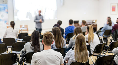 Hörsaal mit besetzten Stühlen. Das Publikum ist von hinten zu sehen. Vor ihnen steht ein Mann im grauen Hemd bei einer Präsentation.