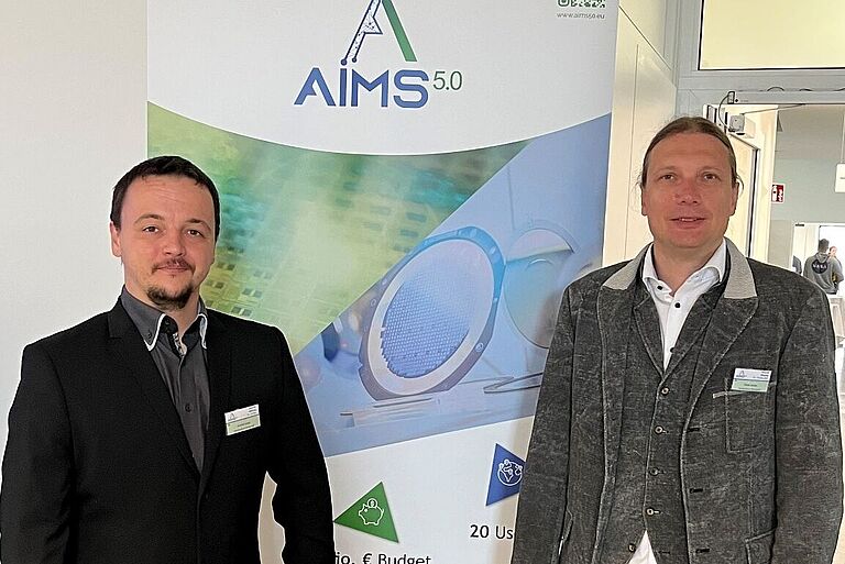 Zwei Männer (Arnold Lange und Prof. Dr. Mark Vehse) stehen vor dem Roll-up-Plakat des Projektes Aim 5.0